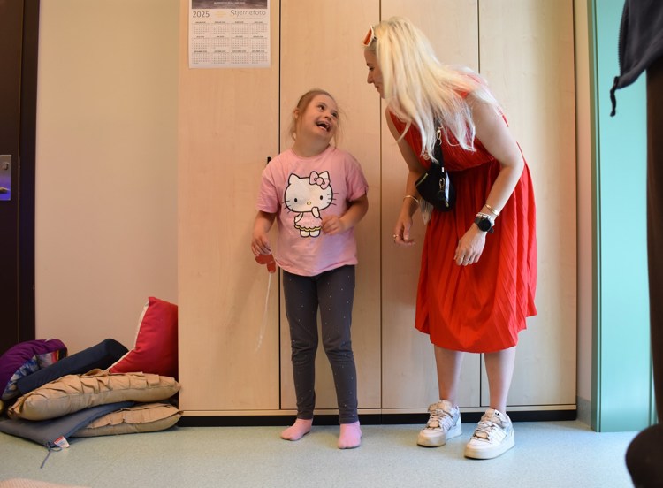 Jente med downs syndrom står med Hello Kitty-genser ved siden av voksen dame i rød kjole og med lyst hår. De smiler til hverandre. 