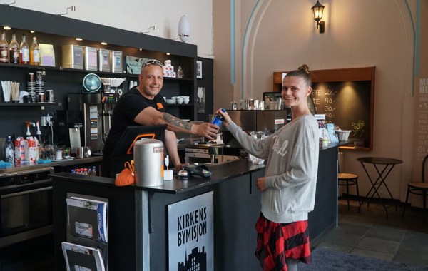 Kafé på Kirkens Bymisjon, en mann står bak disken og serverer en brus til en kvinne på andre siden.